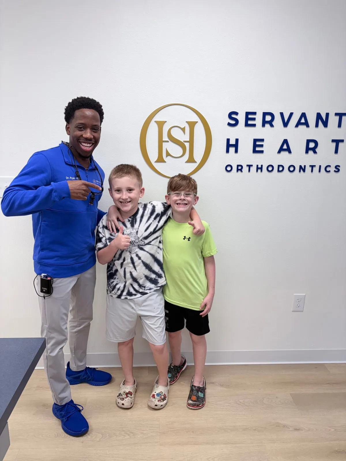 Two young patients with Dr. Ryan Allo in front of the Servant Heart Orthodontics sign in Rowlett, TX